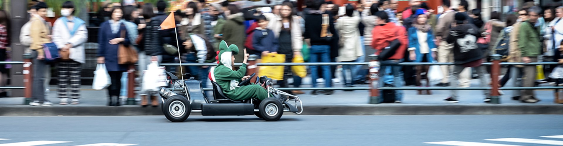 man-in-costume-driving-mariocart-1920x500 A man in a dinosaur costume zipping down a busy Japanese city street in a go-cart with a flag on the back.