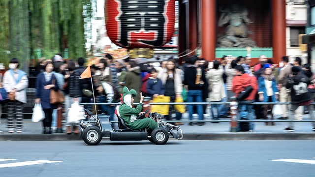 A man in a dinosaur costume zipping down a busy Japanese city street in a go-cart with a flag on the back.
