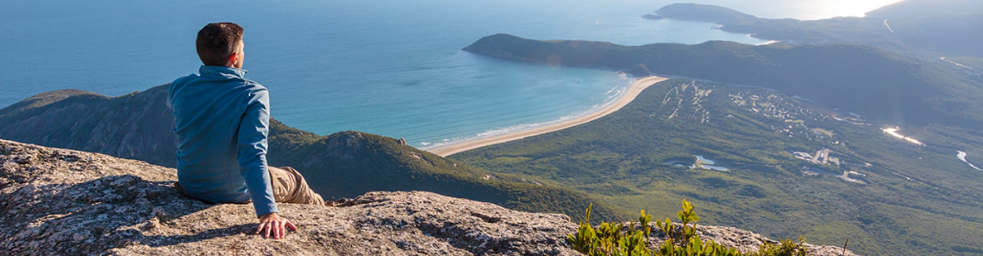 man-on-cliff-over-beach-1920x500 A man sits atop a cliff on a sunny day, leaning back on one hand to overlook a tropical scene of green hills leading to buildings and a beach.