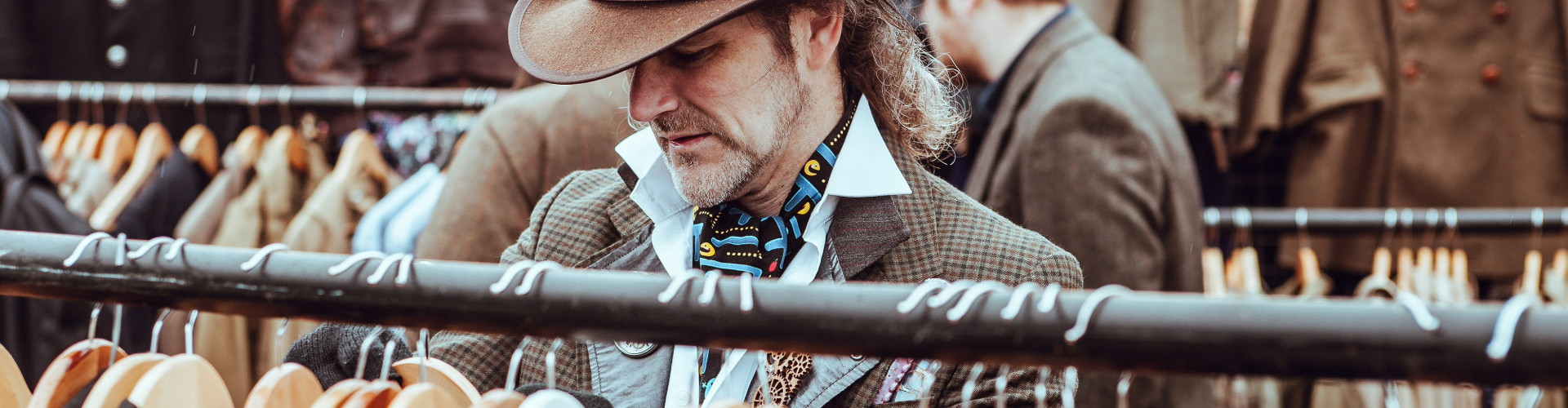 man-shopping-sydney-markets-1920x500 A man in a brimmed hat, popped collar, scarf and jacket, sifts through clothes on a rack at an outdoor market.