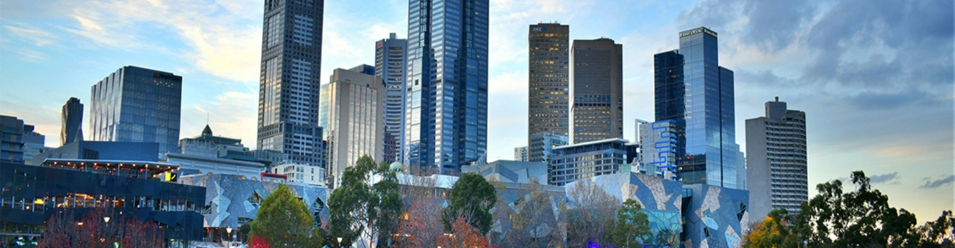 melbourne-city-skyline-vic-1920x500 The Melbourne city's business district skyscrapers, framed by trees and blue sky.