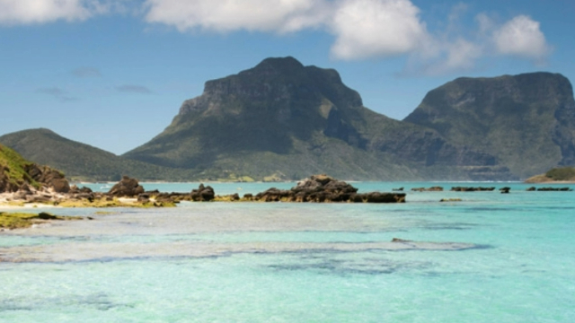 Two treed mountains rising out of turquoise blue ocean into a blue sky, with green covered rocks and small beach in the foreground.