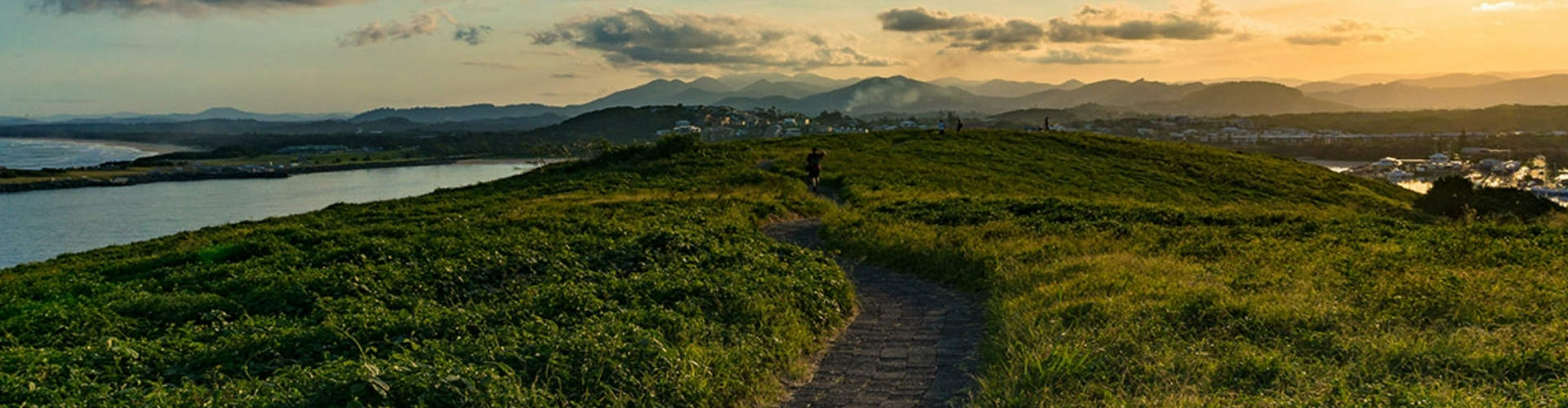muttonbird-island-coffs-harbour-1920x500 A dirt path along the top of an island, lit by a sunset over mountains in the distance.