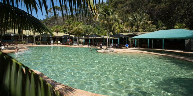  A large turquoise pool at a resort surrounded by blue sky, tropical landscaping, awnings and seating areas. 