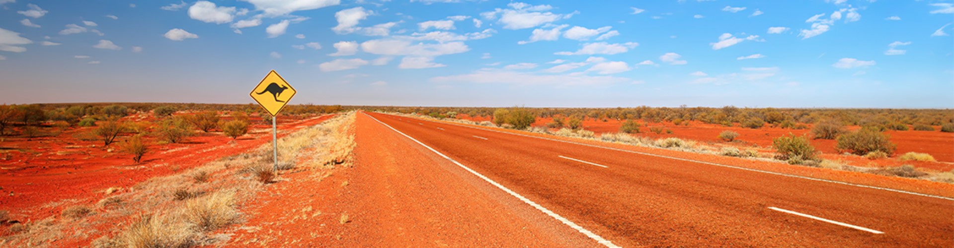 outback-road-with-kangaroo-sign-1920x500 An empty paved road, stained orange from the dusty, scrubby, outback around it and a road sign with a jumping kangaroo, under blue sky.