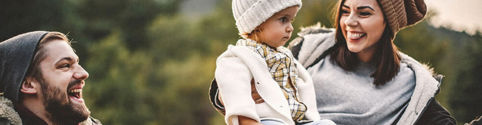 parents-and-baby-in-winter-hats-1920x500 A man, woman and child in winter hats, laughing, with trees behind them.