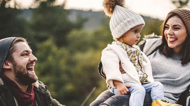 A man, woman and child in winter hats, laughing, with trees behind them. 