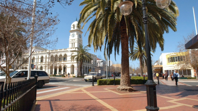 People walking around a town square with a historic clock tower opposite a sunny park with large palm trees.