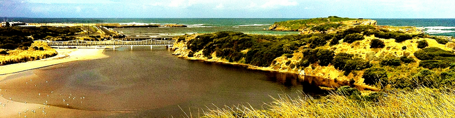 pickering-point-warrnambool-vic-1920x500 A wide view of a lagoon created by two grassy heads, connected by a wooden bridge leading out to the sea.