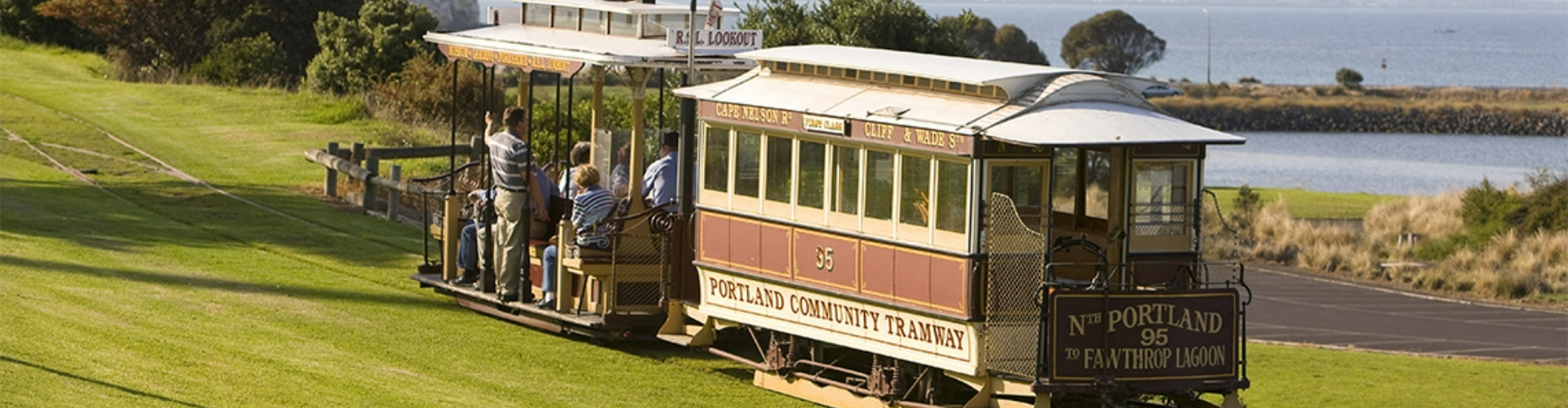 portland-cable-tram-vic-1920x500 A historic tram with two cars, one closed off with windows, and one with a roof and seats, but no walls, going through a sunny green park by the sea.