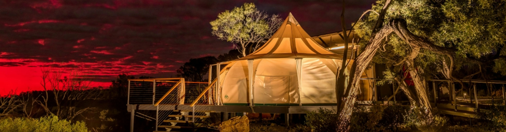 premade-camping-tent-1920x500 A large canvas tent on a wooden verandah at sunset, surrounded by gum trees.