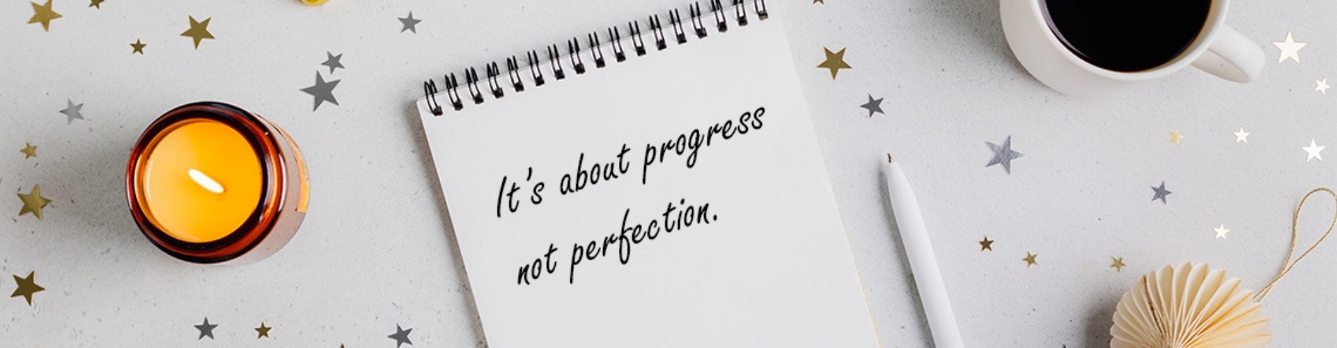 Resolutions 2024 Notepad with writing that says It's about progress not perfection, next to candle and coffee and gold balloon numbers 2024 viewed from above