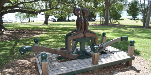 A wooden sculpture and teeter-totter for kids in a sunny park, representing a man and a woman facing opposite directions.