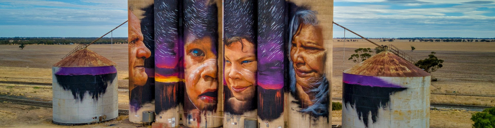 silo-art-sheep-hills-vic-1920x500 Four tall silo towers with a mural spread out across them of a black starry sky over purple sunset, with a man and a woman's face on the two outside silos, and two children's faces on the middle towers.