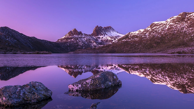 Snow capped mountain peaks backlit in bright purple by a sunset, and reflected in a lake below.