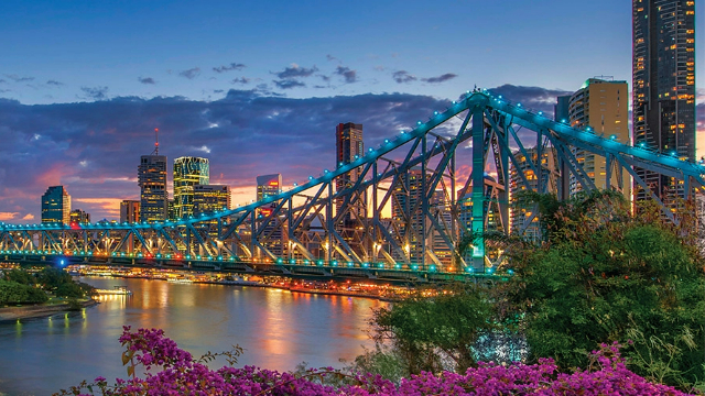 A double-steepled bridge, with girders lit with blue lights over a river reflecting glowing city skyscraper lights.