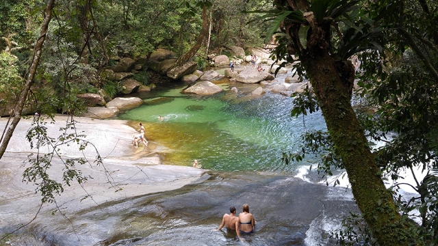 man and woman in swim suits sit in shallow water on smooth rock at peaceful rocky pool surrounded by rainforest while children swim and play in the water