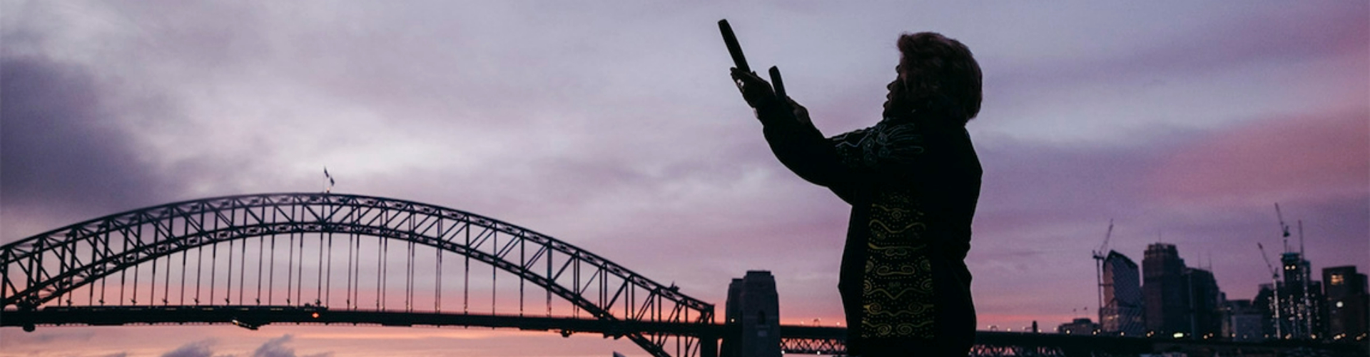 sydney-aboriginal-dreaming-tour-nsw-1920x500 Aunty Margret, an Aboriginal woman, storytelling at the Sydney Harbour shore, silhouetted by a sunrise.