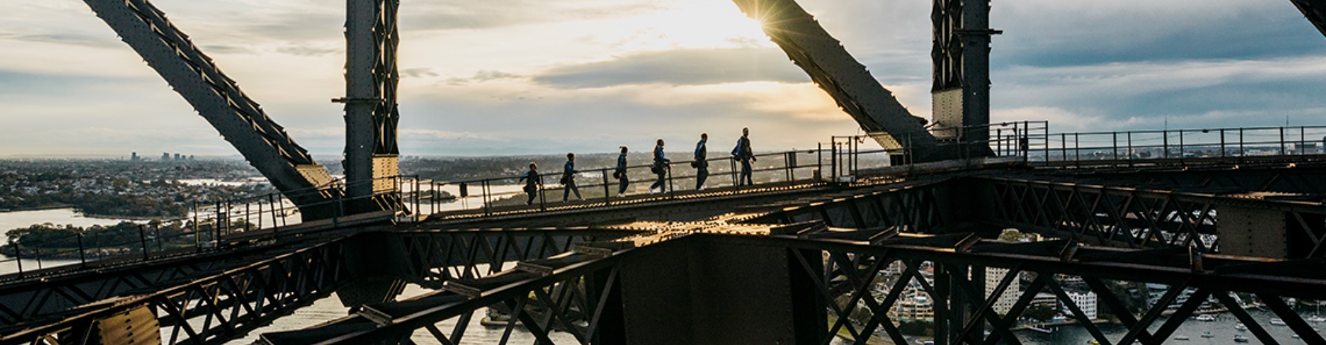 sydney-bridge-climb-burrawa-nsw-1920x500 Six people walk along the gridwork at the top of the Sydney Harbour Bridge at sunrise.