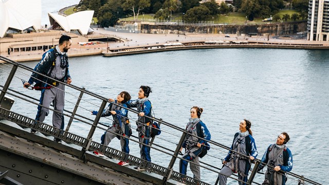 A man leads a family up the Sydney Harbour Bridge girders, with the harbour and Opera house below. 