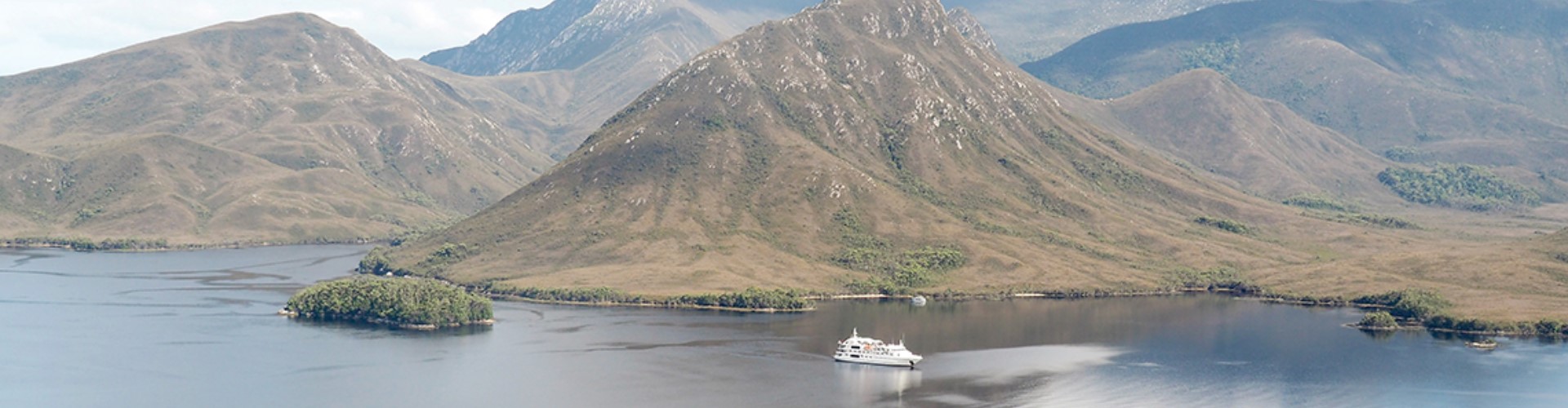 tasmania-cruise-boat-1920x500 A wide vista of two near-mountainous pointed hills rising above a waterway where a white Coral expeditions cruise boat glides along in front of the tallest mountain.