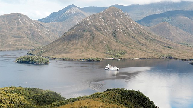 A wide vista of two near-mountainous pointed hills rising above a waterway where a white Coral expeditions cruise boat glides along in front of the tallest mountain.