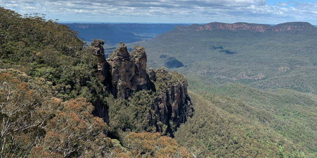 Three spires of rock rise out of a heavily treed mountain hillside, with hills and mountains stretching into the distance. 