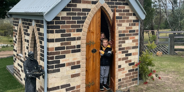 Two children peek out from the wooden door of a very small brick church set on grass near a paddock.