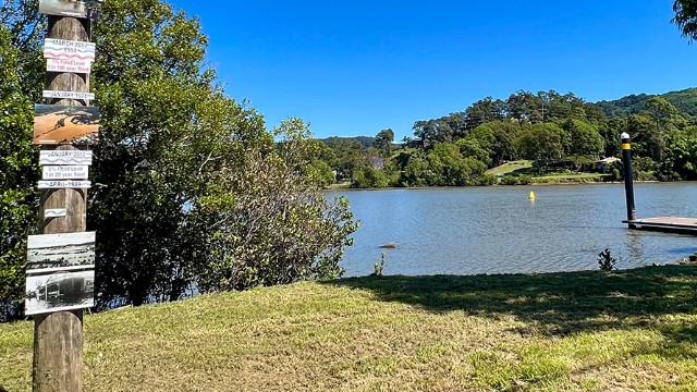 A sunny, treed riverbank, with a pier and a wood post on shore with historical information, photos and swim warnings.
