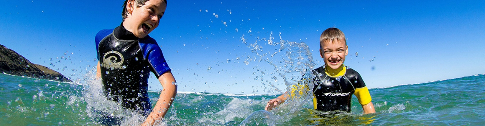 two-boys-swimming-and-laughing-1920x500 Two boys in rash guard shirts splash each other and laugh in waist deep ocean water, under blue sky.