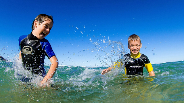 two-boys-swimming-and-laughing-640x360 Two boys in rash guard shirts splash each other and laugh in waist deep ocean water, under blue sky.