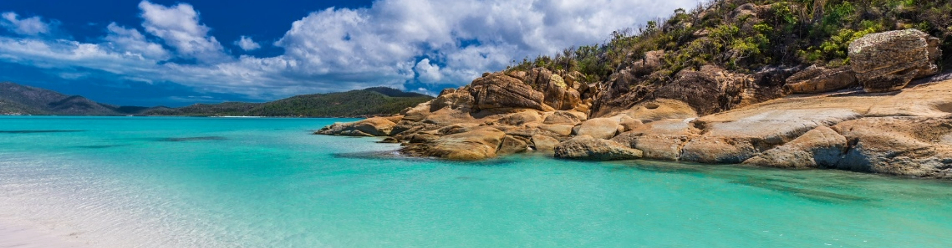 whitehaven-beach-shore-qld-1920x500 A bay of turquoise blue water framed by a rocky outcrop, white sand beach and green hills on the far shore.