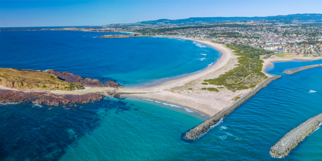 An aerial view of a large, sunny bay with Shellharbour township tucked into a sandy beach coastline that leads leading to a sand peninsula connected to a small island..
