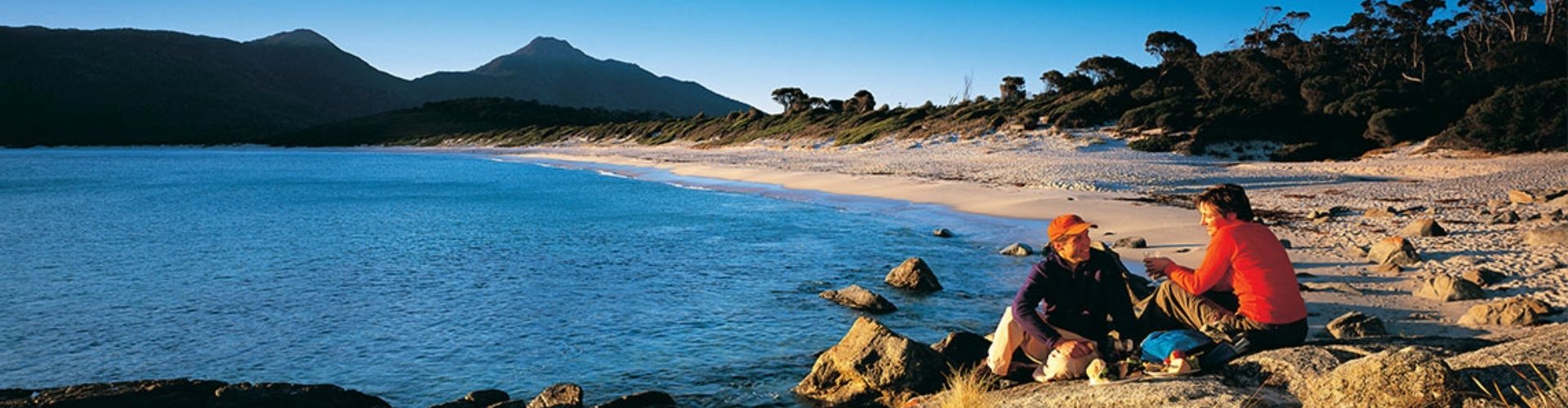 Wineglass Bay TAS couple couple having a picnic sat on the banks of a deserted beach with mountains in the background in the late afternoon