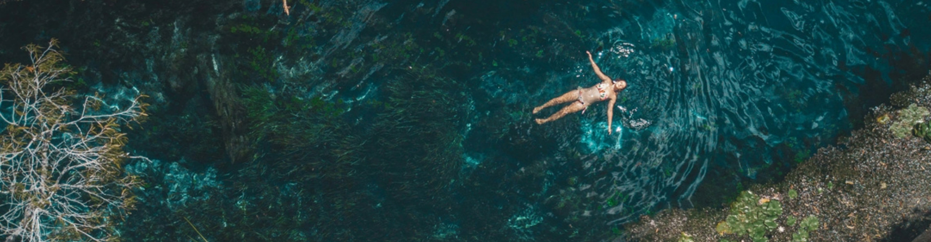 woman-floating-in-lake-1920x500 Aerial view of a woman in a floral bikini lit by the sun as she floats on her back in turquoise water.