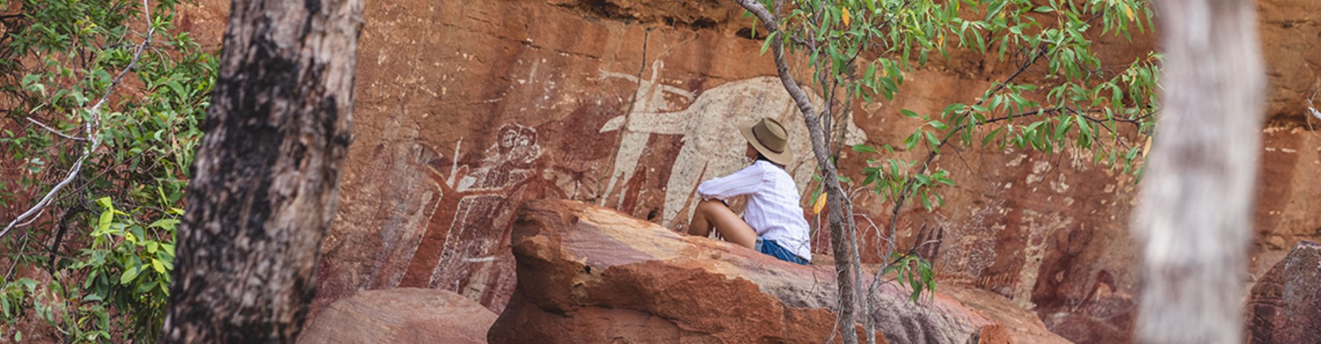 jpeg A woman perches among red rocks and trees, looking at indigenous rock painting of an a human and large animal.