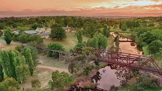 A village nestled among trees near a riverbend with a large rail bridge, and walking bridge further down, lit by a sunset.