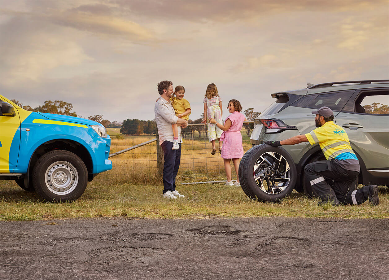 NRMA Roadside assistance staff member helping to replace tyre for vehicle on the side of the road while family of four look on