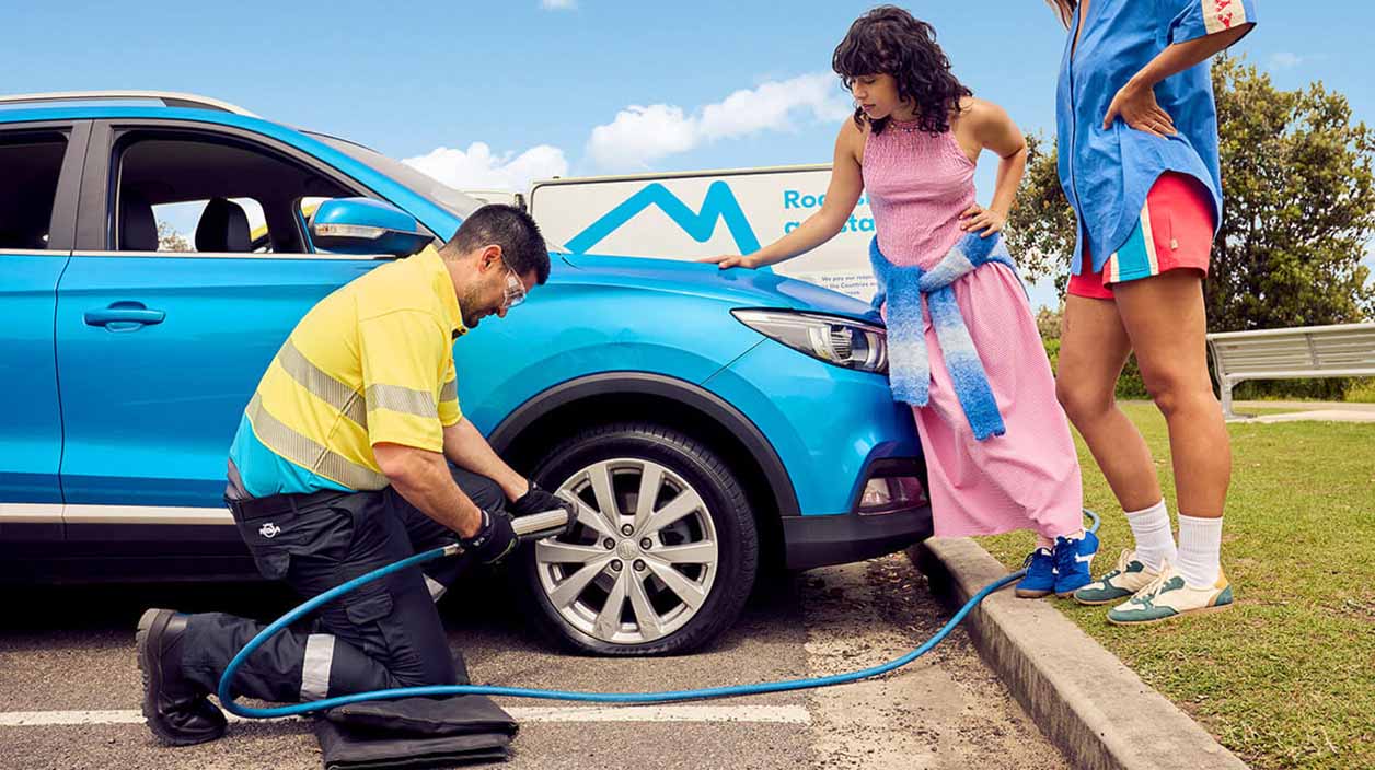 Roadside assistance staff member in yellow and blue uniform kneeling to fix a car tire while two women stand near the front of the car watching