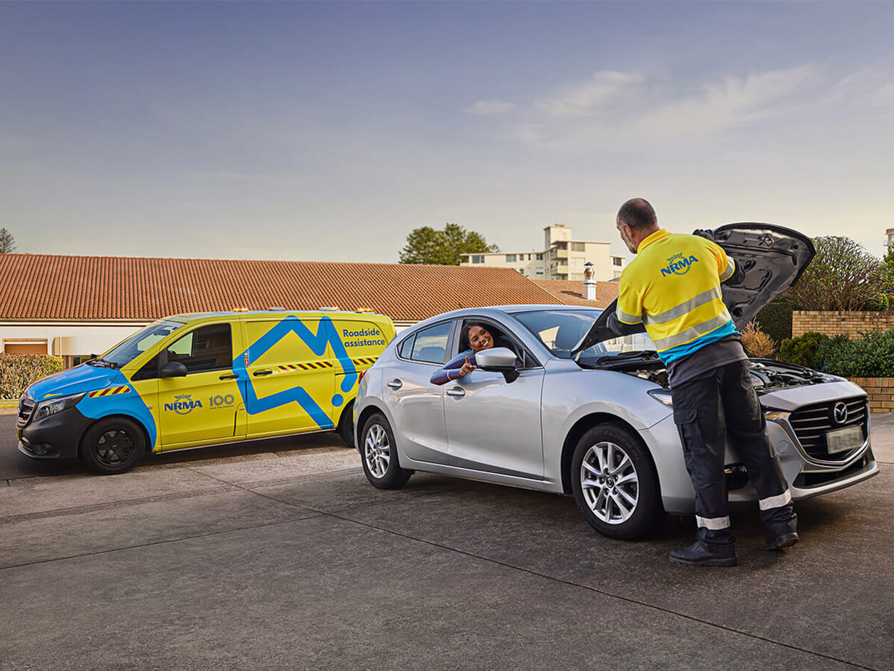 Man in yellow NRMA uniform repairing a car while engaging with a customer sitting in their car