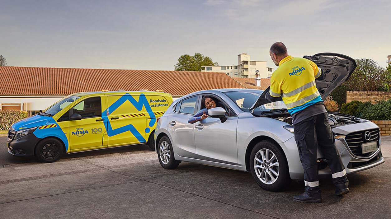 Man in yellow NRMA uniform repairing a car while engaging with a customer sitting in their car