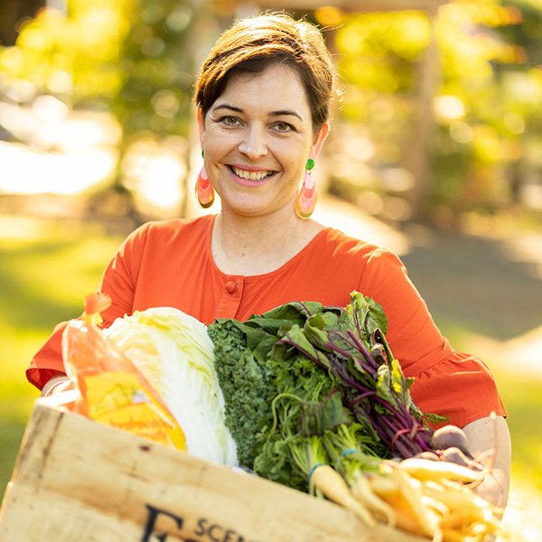 smiling woman with brunette hair wearing an orange shirt and earrings that is holding a box of produce